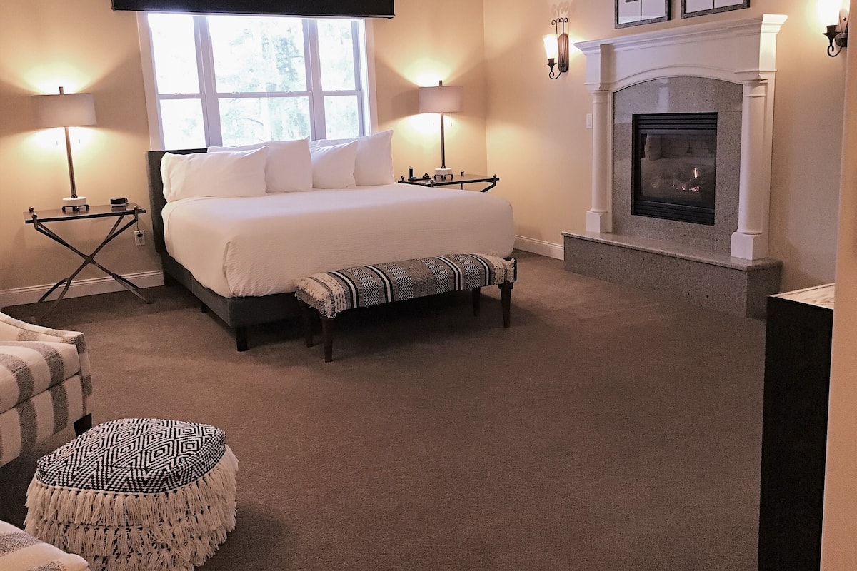 A bedroom with a large bed, bedside lamps, a fireplace, and a window with blinds.