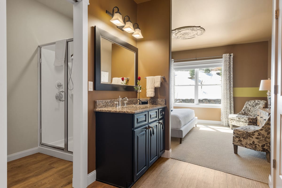A bathroom with a glass shower, dark cabinets, a large mirror, and a view of a bedroom with a bed and a chair.