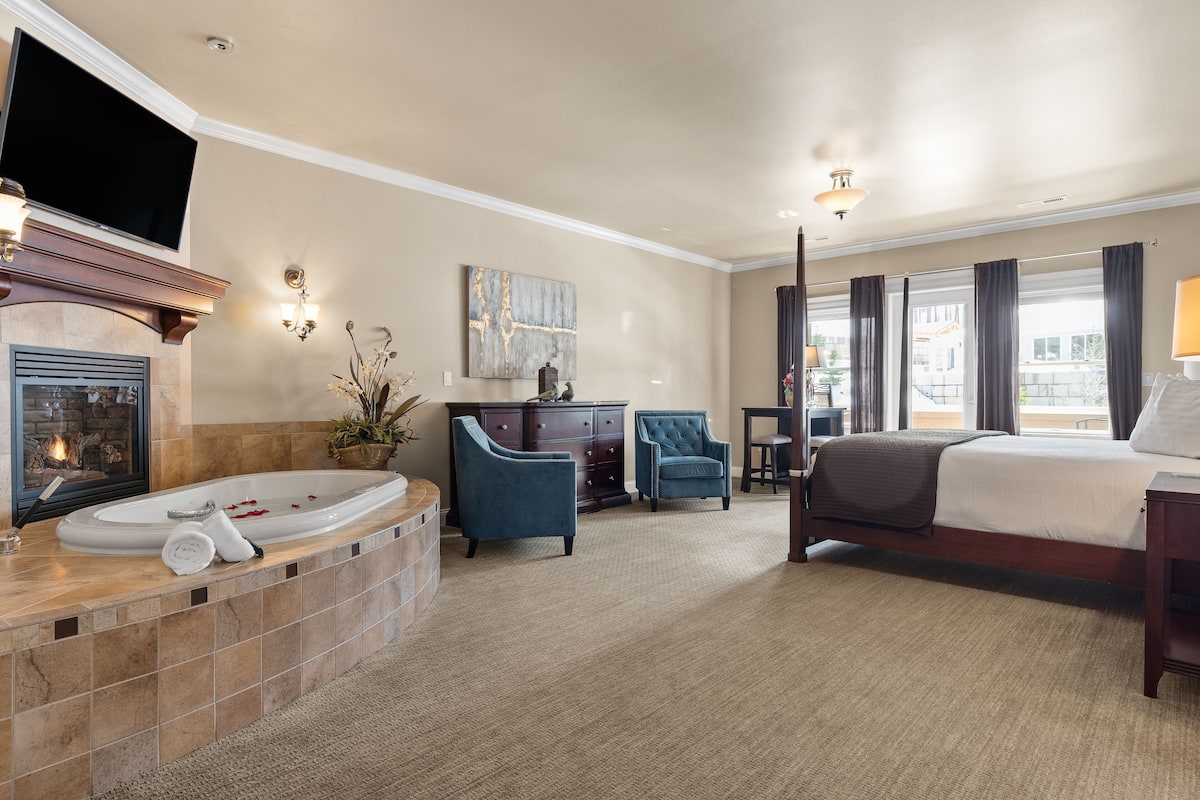 A hotel room with a large bed, a sitting area with a chair and a table, a fireplace, and a bathtub.