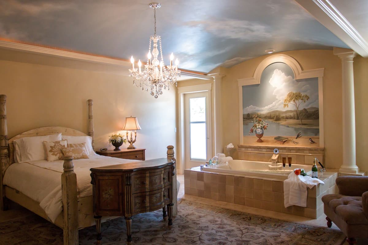 A bedroom with a large bed, a wooden dresser, a chandelier, a nightstand, and a bathroom with a bathtub, mirror, and towel rack.