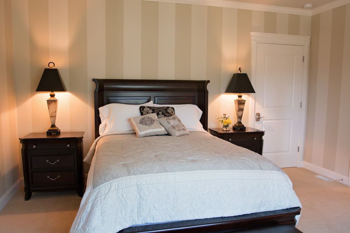A neatly made bed with a white comforter and decorative pillows, flanked by two bedside tables with lamps, in a room with striped wallpaper and a closed door.