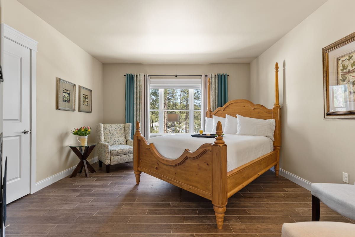 A bedroom with a wooden four-poster bed, a chair, a small table with a plant, and a window with curtains.