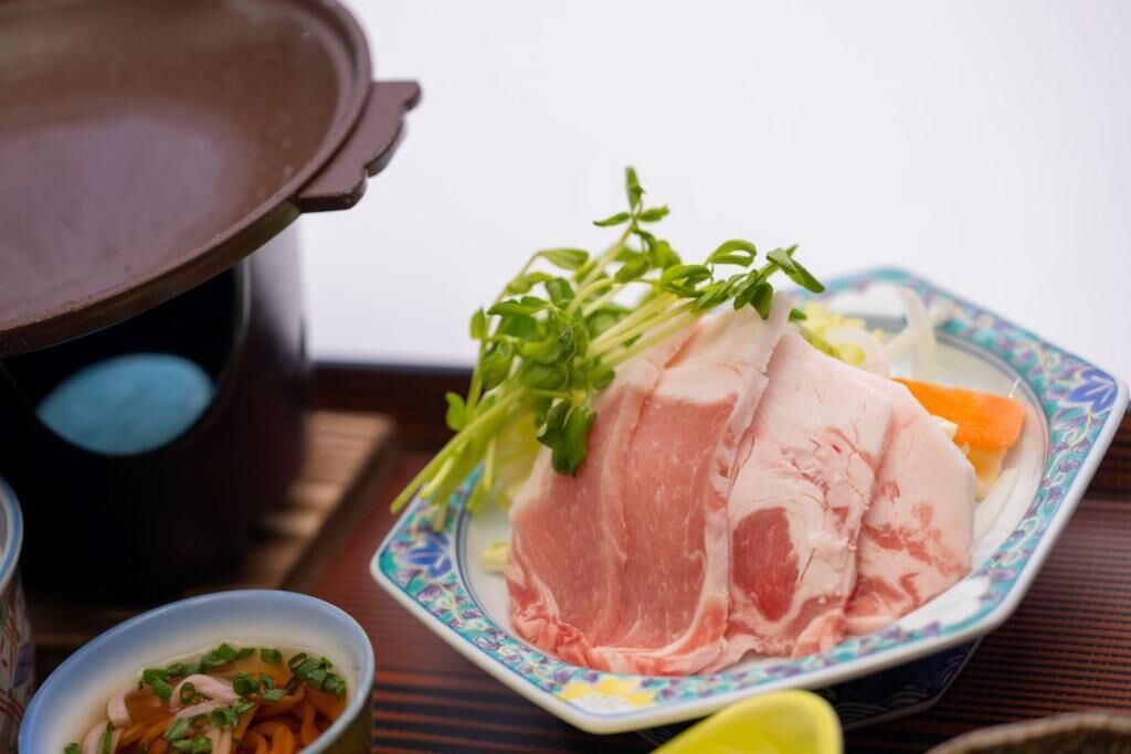 Una preparación tradicional japonesa de olla caliente, con una olla de cerámica, un plato con carne y verduras en rodajas, y un tazón pequeño de sopa.