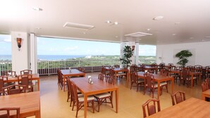 Un vaste espace dédié à la salle à manger, avec de nombreuses tables et chaises en bois, de grandes fenêtres offrant une vue sur la mer et les bâtiments lointains, et un balcon doté d’une balustrade.