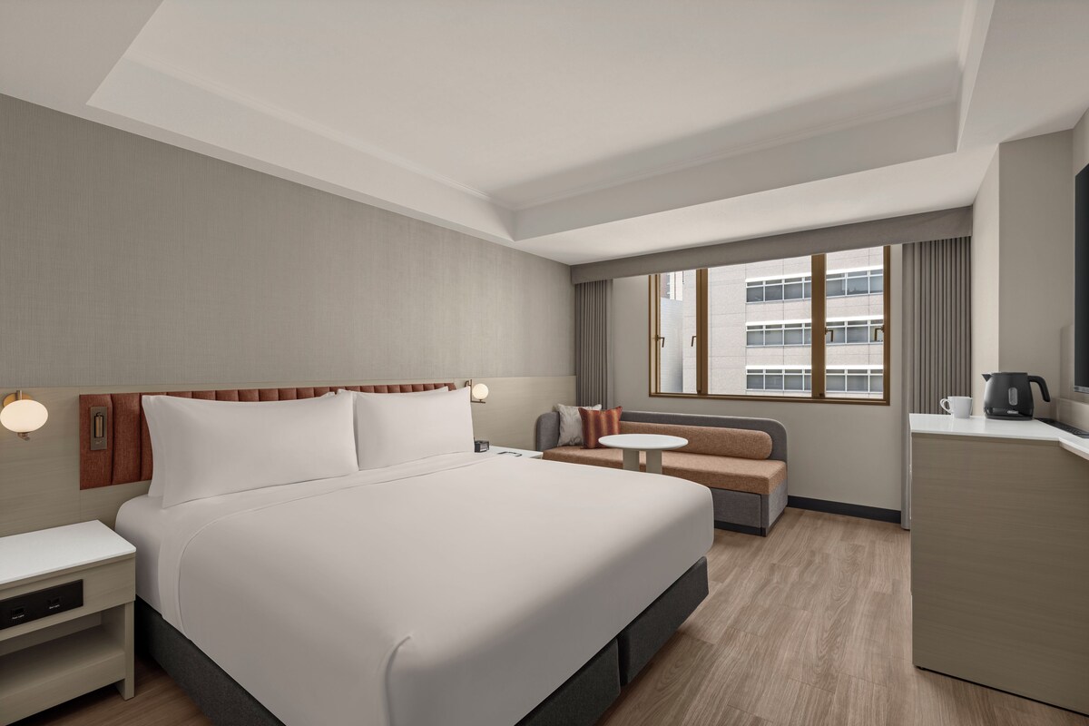 Ein Hotelzimmer mit Bett, Nachttisch, Lampe, Sofa und einem kleinen Tisch mit Wasserkocher.