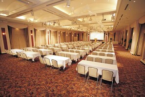 Un salón de conferencias con filas de mesas y sillas, piso alfombrado y una pantalla grande al frente.