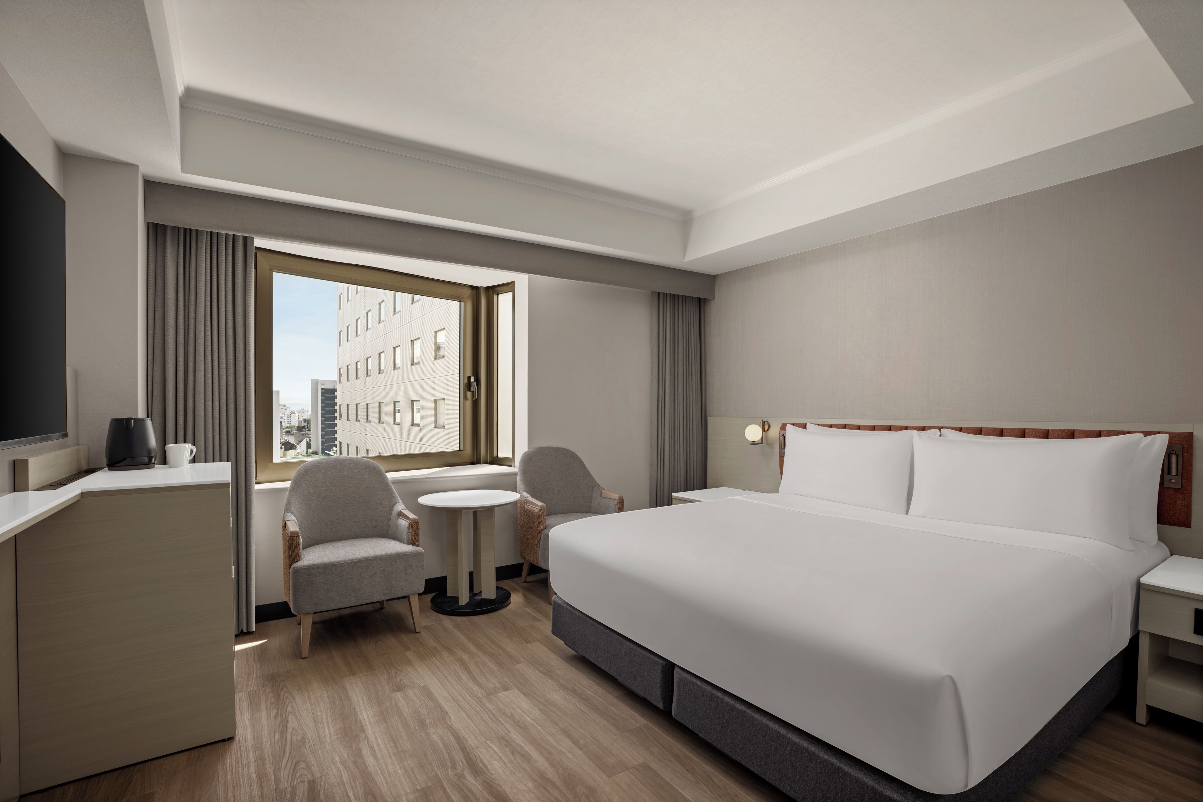 A hotel room with a large bed, a desk, a chair, a small table, and a view of the city through the window.