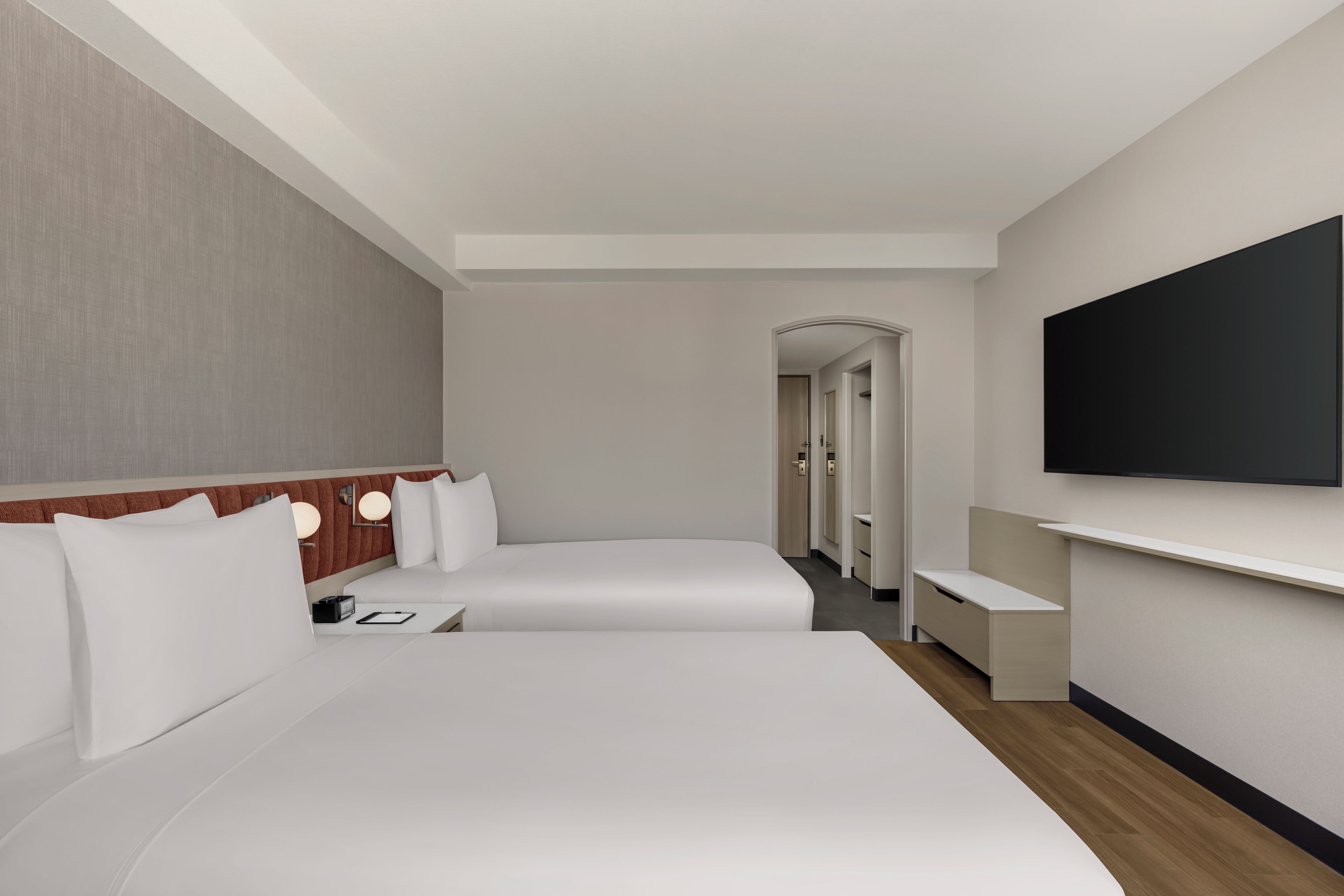 A hotel room with two beds, a flat-screen TV, a wooden headboard, and a hallway leading to other rooms.
