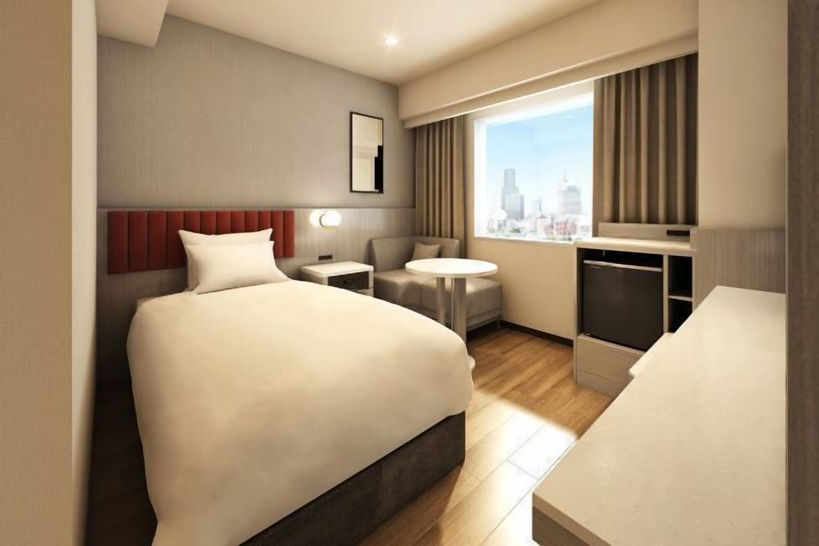 Ein Hotelzimmer mit einem großen Bett, einem Schreibtisch, einem Sessel, einem kleinen Tisch und einem Fenster mit Blick auf die Stadt.