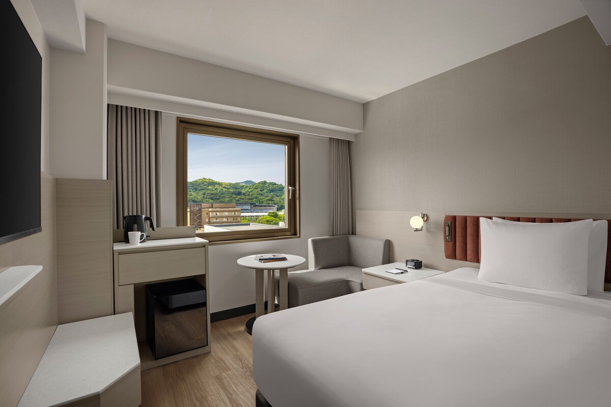 Une chambre d’hôtel comprenant un lit, un bureau, une chaise, une petite table et une fenêtre donnant sur un paysage verdoyant et des bâtiments.