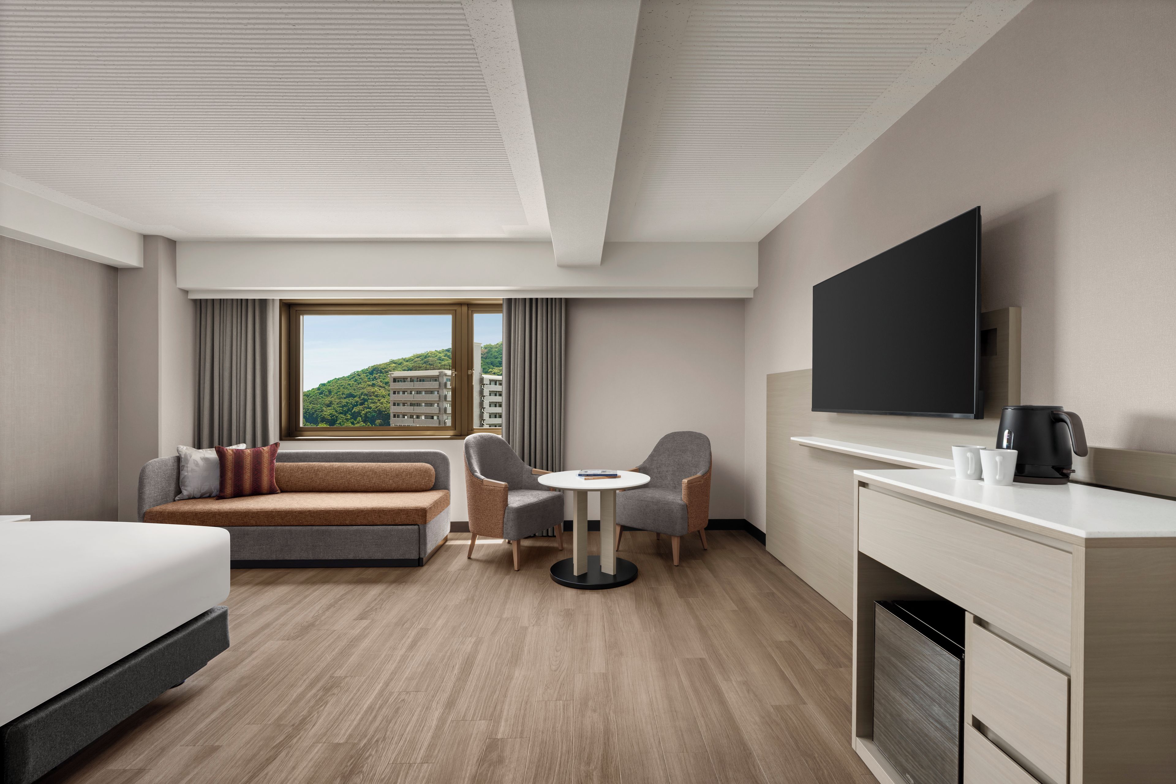 A hotel room with a bed, a sofa, a dining table, two chairs, a TV, a coffee maker, and a view of a building through the window.