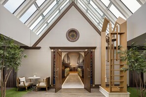 Un intérieur moderne avec un plafond haut et des puits de lumière, une large ouverture centrale donnant sur un espace de salle à manger, et une structure en bois surmontée d’une cloche, située à droite.