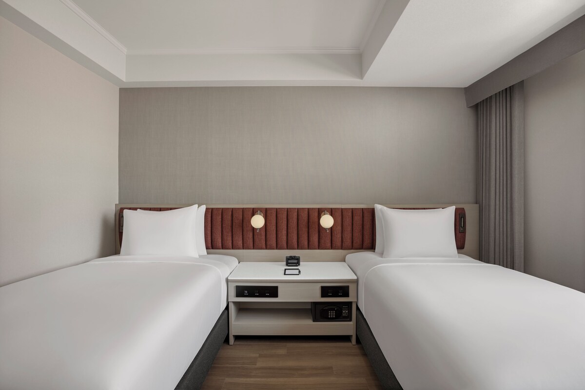 Ein Hotelzimmer mit zwei Einzelbetten, einem Nachttisch mit Lampe und einer Bedieneinheit für die Zimmerausstattung.