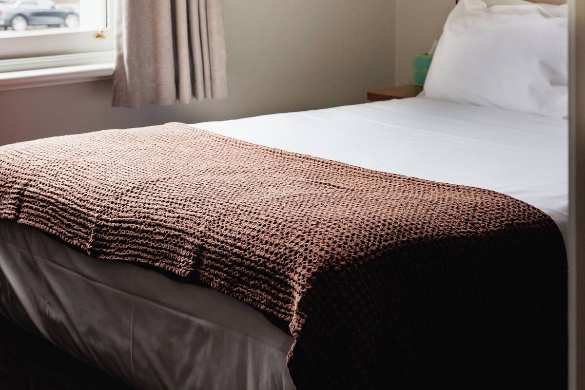 Habitación de hotel con cama, un cuadro en la pared, una ventana con cortinas y una mesita de noche con lámpara.