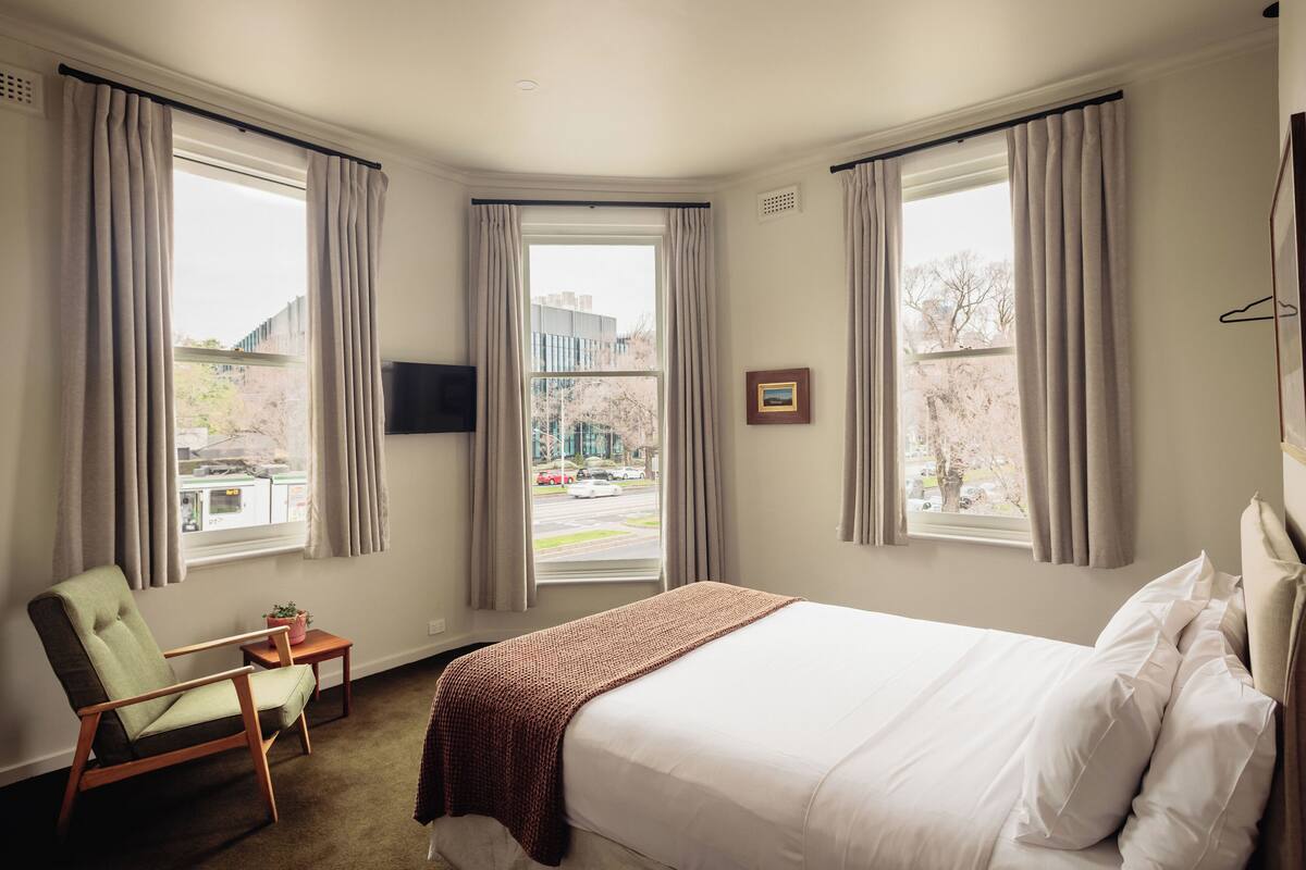 Una habitación de hotel con cama, una silla, una mesita y dos ventanas con cortinas.