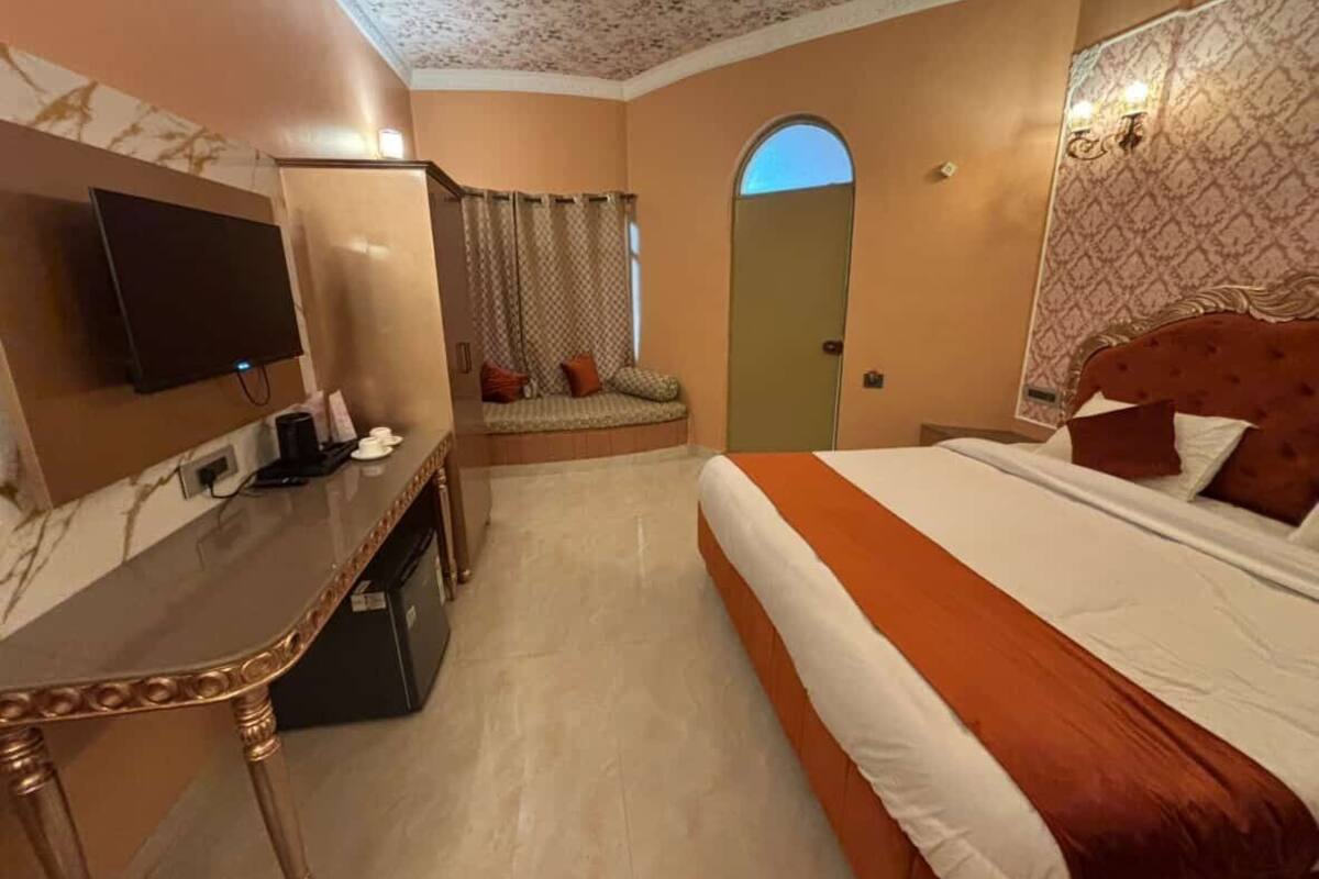 A hotel room with a bed, a desk with a flat-screen TV, a seating area with a sofa and curtains, and a patterned wall.
