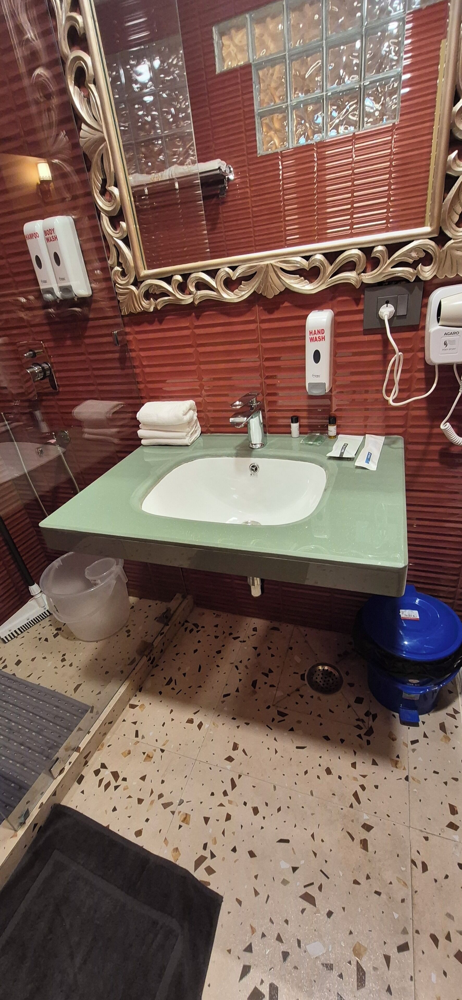 Un baño con un lavamanos verde, un espejo con un marco dorado ornamentado y una pared de azulejos rojos.