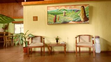A room with two wooden chairs, a small table, a potted plant, and a painting of a landscape on the wall.