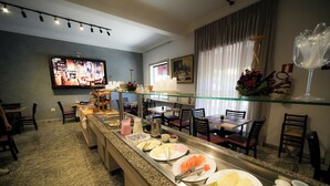 A buffet setup with various food items and utensils, a television displaying a restaurant scene, and a dining area with chairs and tables. - Hotel Euro Suíte Poços de Caldas By Nacional Inn (Poços de Caldas)