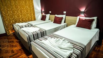 Quarto de hotel com três camas de solteiro, cada uma com uma colcha branca e um travesseiro vermelho. O quarto possui piso de madeira e uma cortina estampada em uma das paredes.