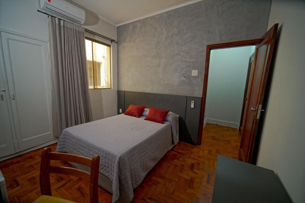 Quarto com cama, uma cadeira, escrivaninha, ar condicionado e janela com cortinas.