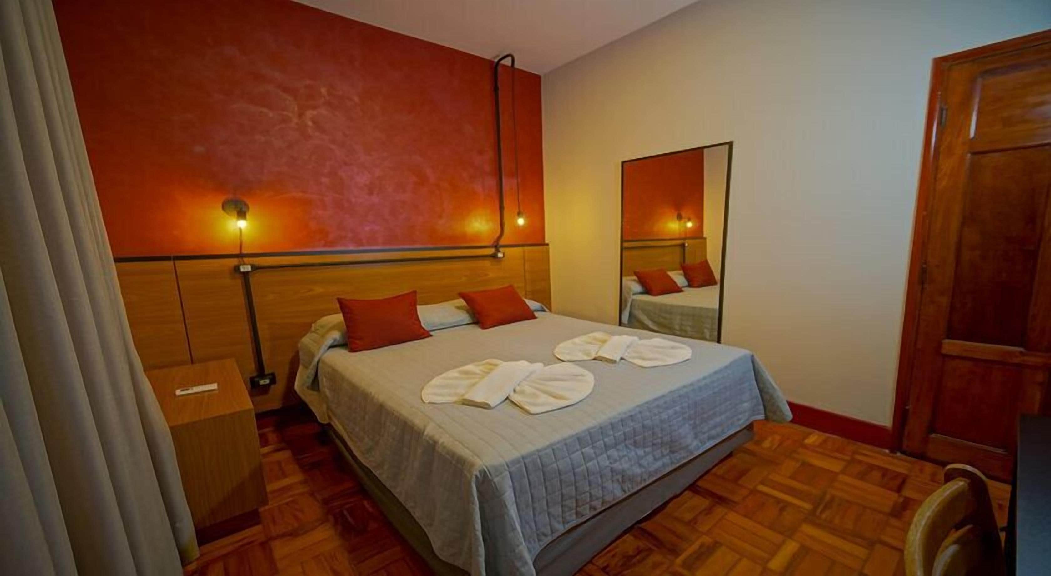 Quarto de hotel com uma cama grande, cabeceira de madeira, mesa de cabeceira, espelho e piso de madeira.