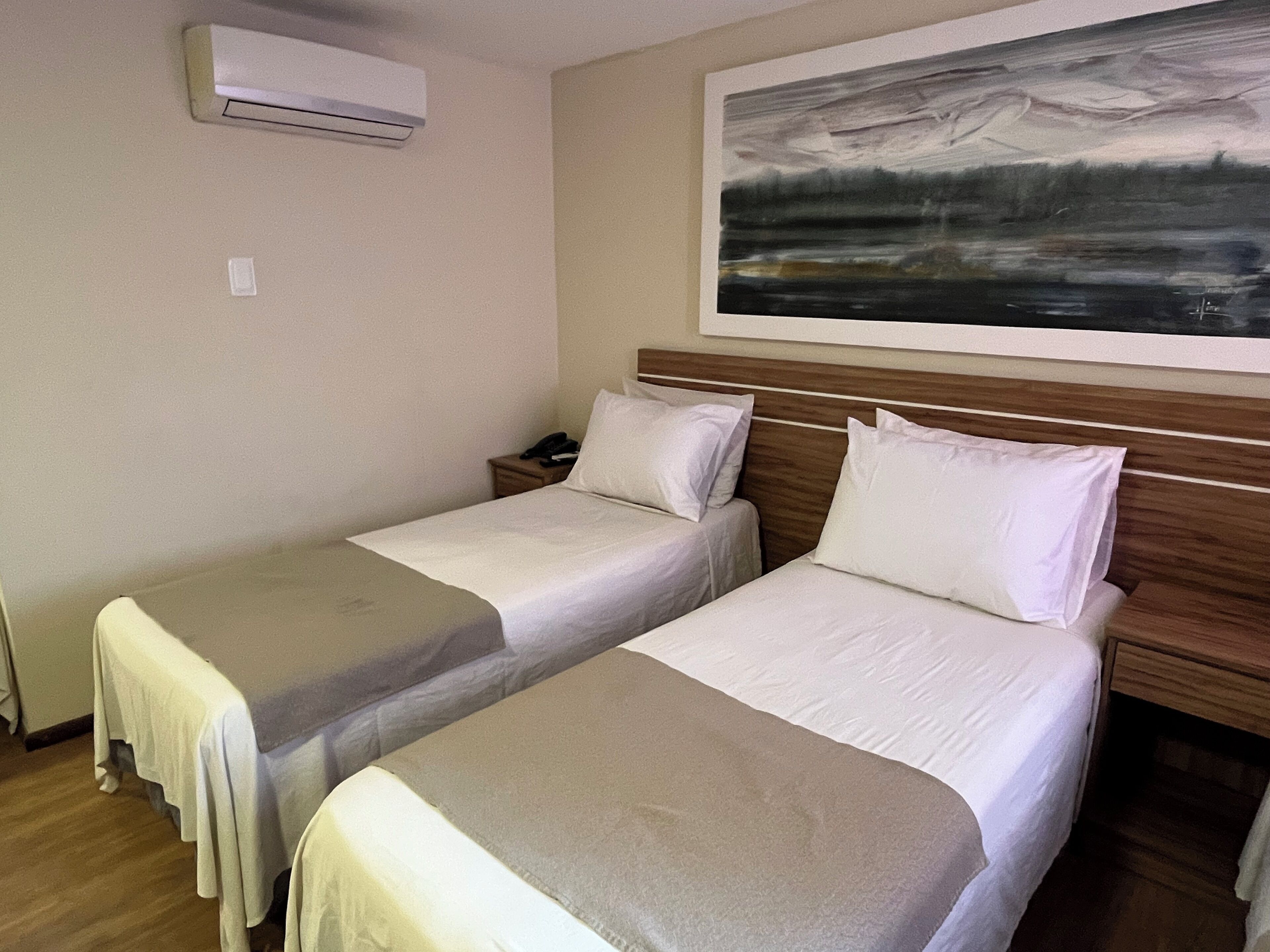 A hotel room with two beds, a painting on the wall, and an air conditioning unit.