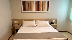 Une chambre d’hôtel avec un grand lit, deux tables de chevet, une petite table, un téléphone fixé au mur et un tableau au-dessus du lit.