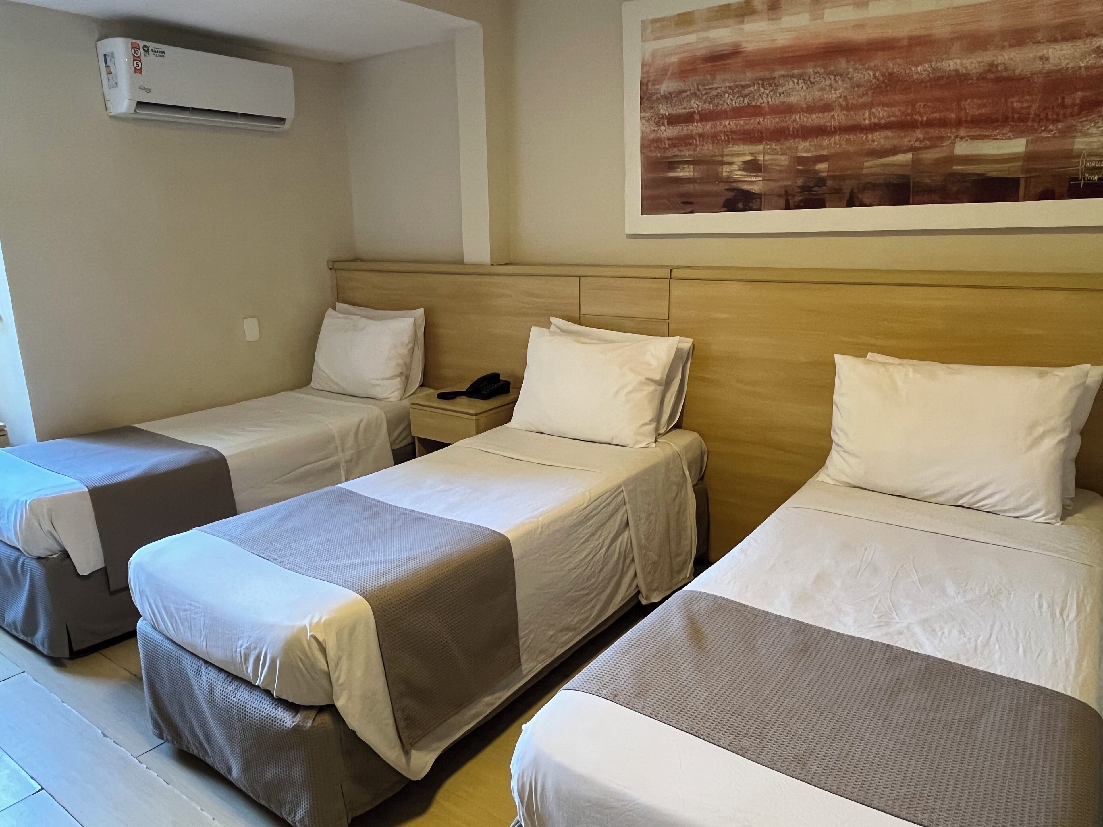 Quarto de hotel com três camas individuais, telefone ao lado da cama e ar condicionado na parede.