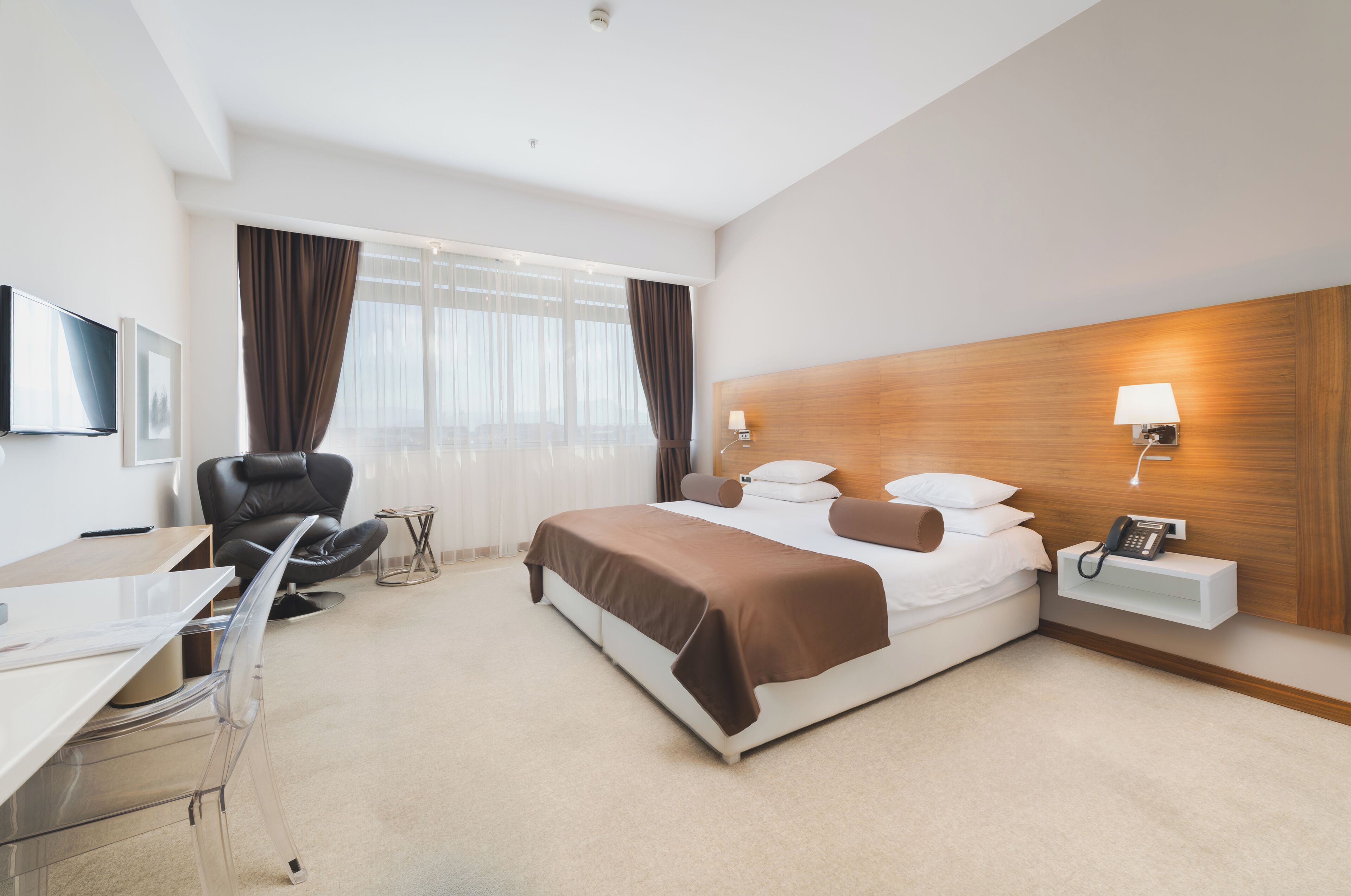 A hotel room with a bed, desk, chair, and a bedside table with a lamp.