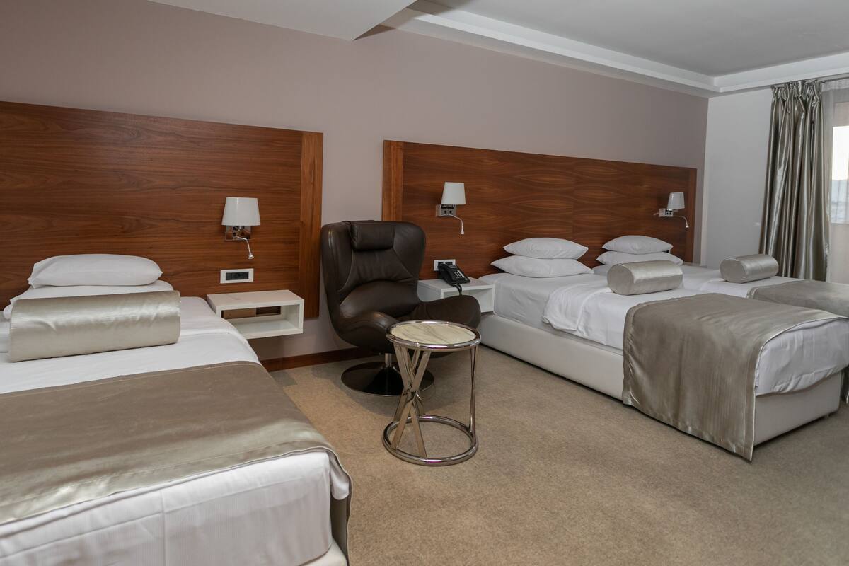 Una habitación de hotel con dos camas individuales, una cama doble, un sillón de cuero, una mesita redonda y dos lámparas de noche.