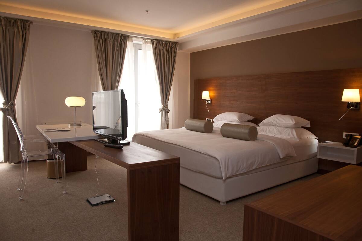 Una habitación de hotel con una cama grande, lámparas de noche, un escritorio con televisor y un cesto de basura.