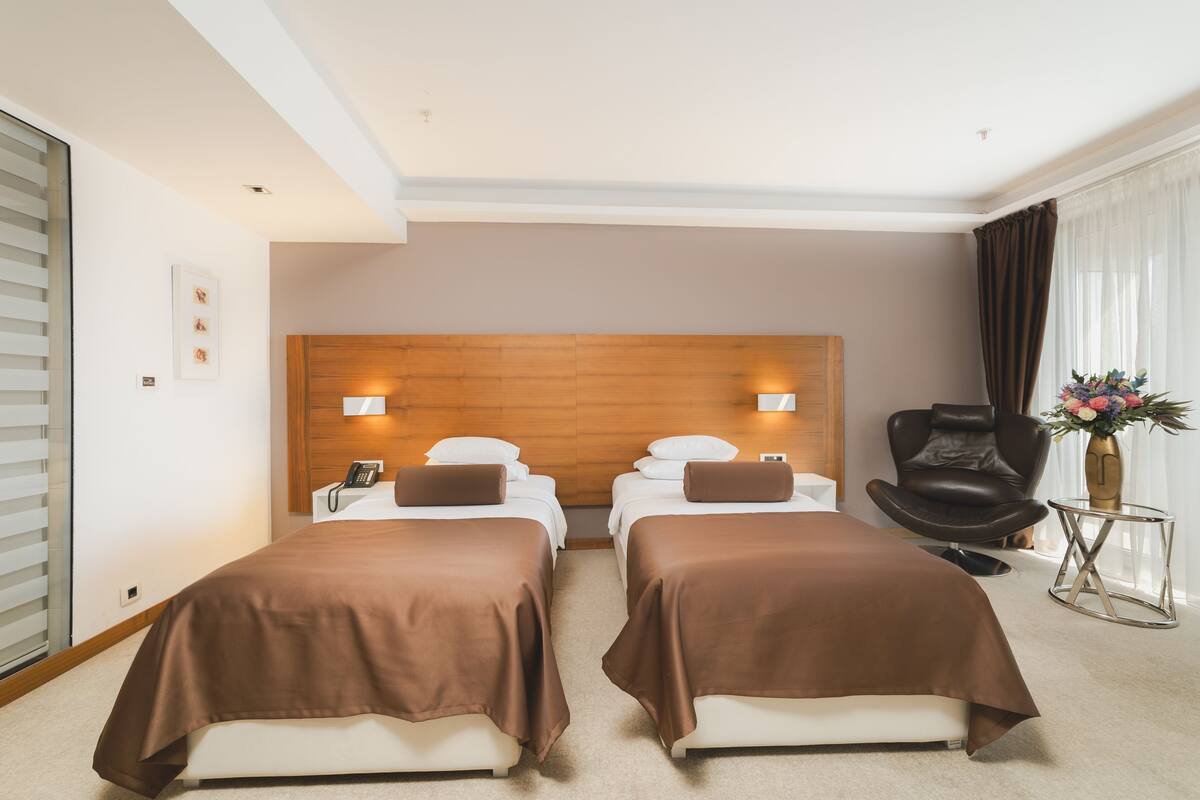 Habitación de hotel con dos camas, cabecero de madera, lámparas de noche, un sillón de cuero, una mesita y un jarrón con flores.