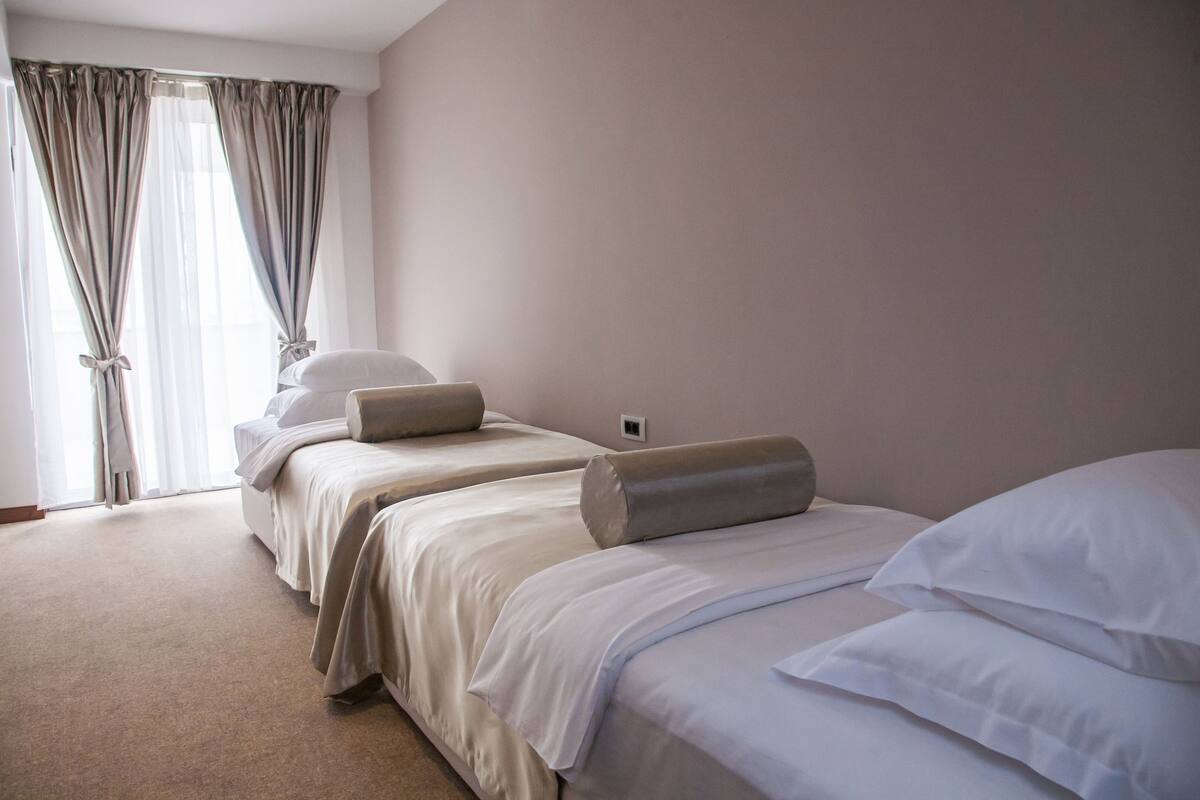 Una habitación de hotel con dos camas, ropa de cama color beige y un ventanal con cortinas.
