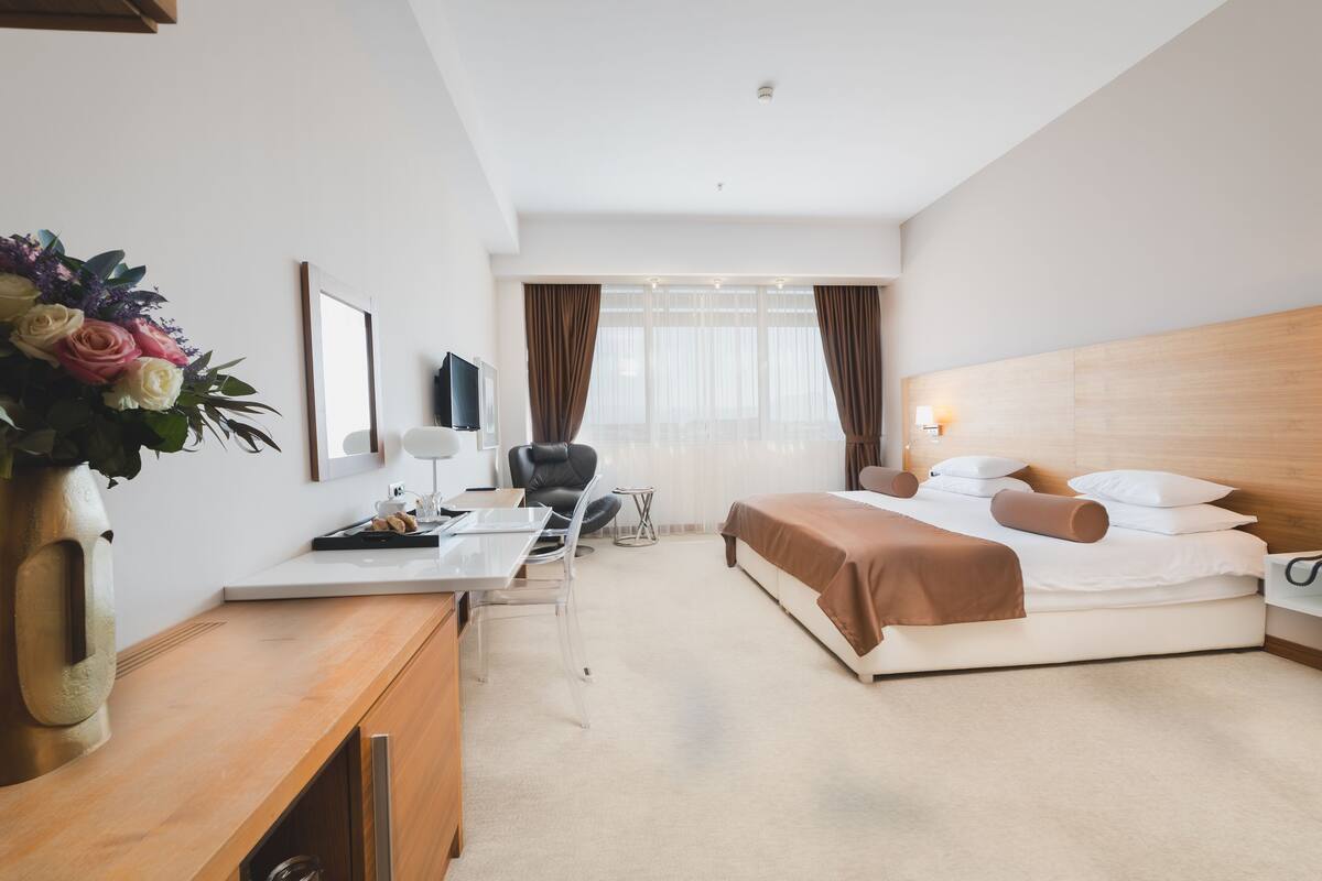 Una habitación de hotel moderna con cama, mesita de noche, silla, escritorio y un gran ventanal con cortinas.