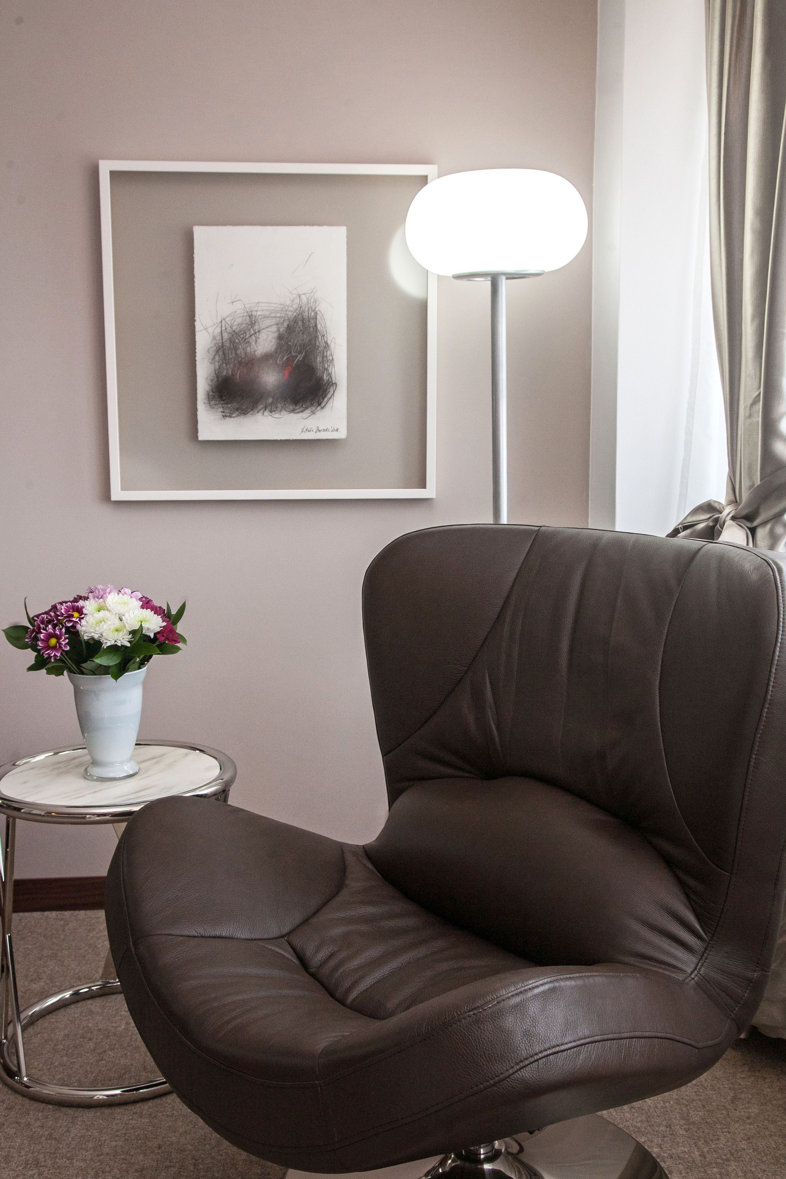 A brown leather chair, a small white vase with flowers, a framed abstract painting, and a floor lamp.