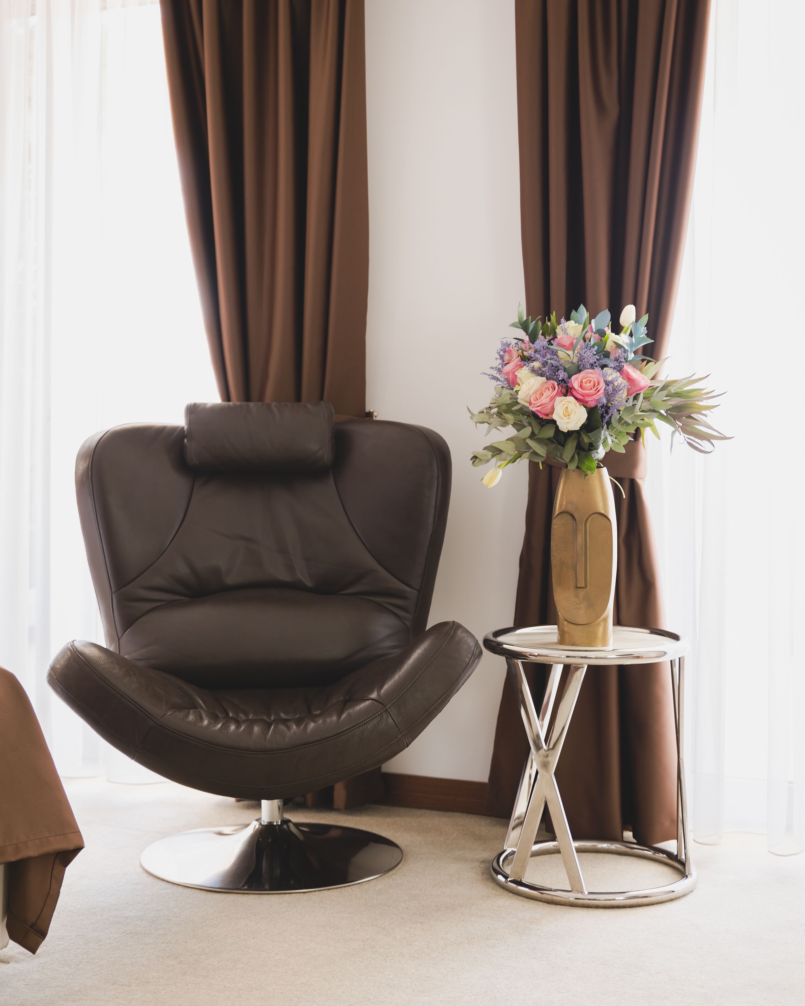 Une chambre moderne avec un fauteuil en cuir, une petite table ronde ornée d’un vase de fleurs et des rideaux.