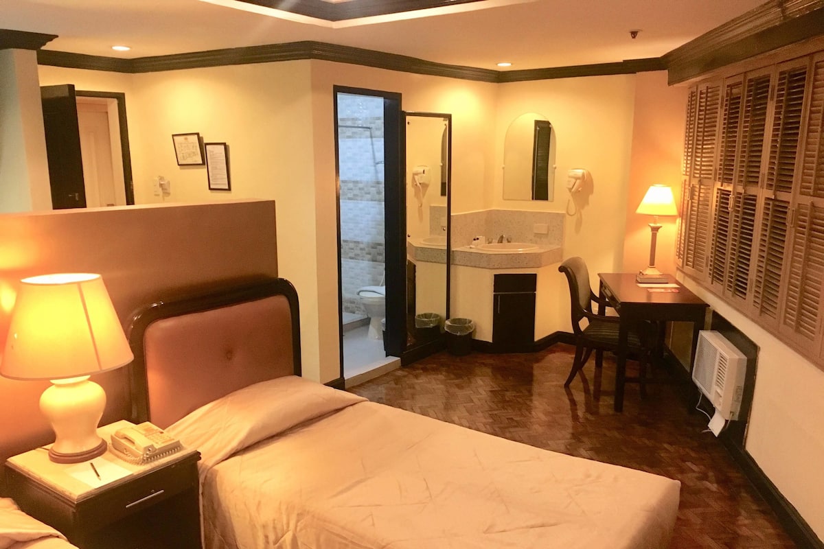 Habitación de hotel con cama, mesita de noche con lámpara, escritorio con silla, espejo y ventana con persianas.