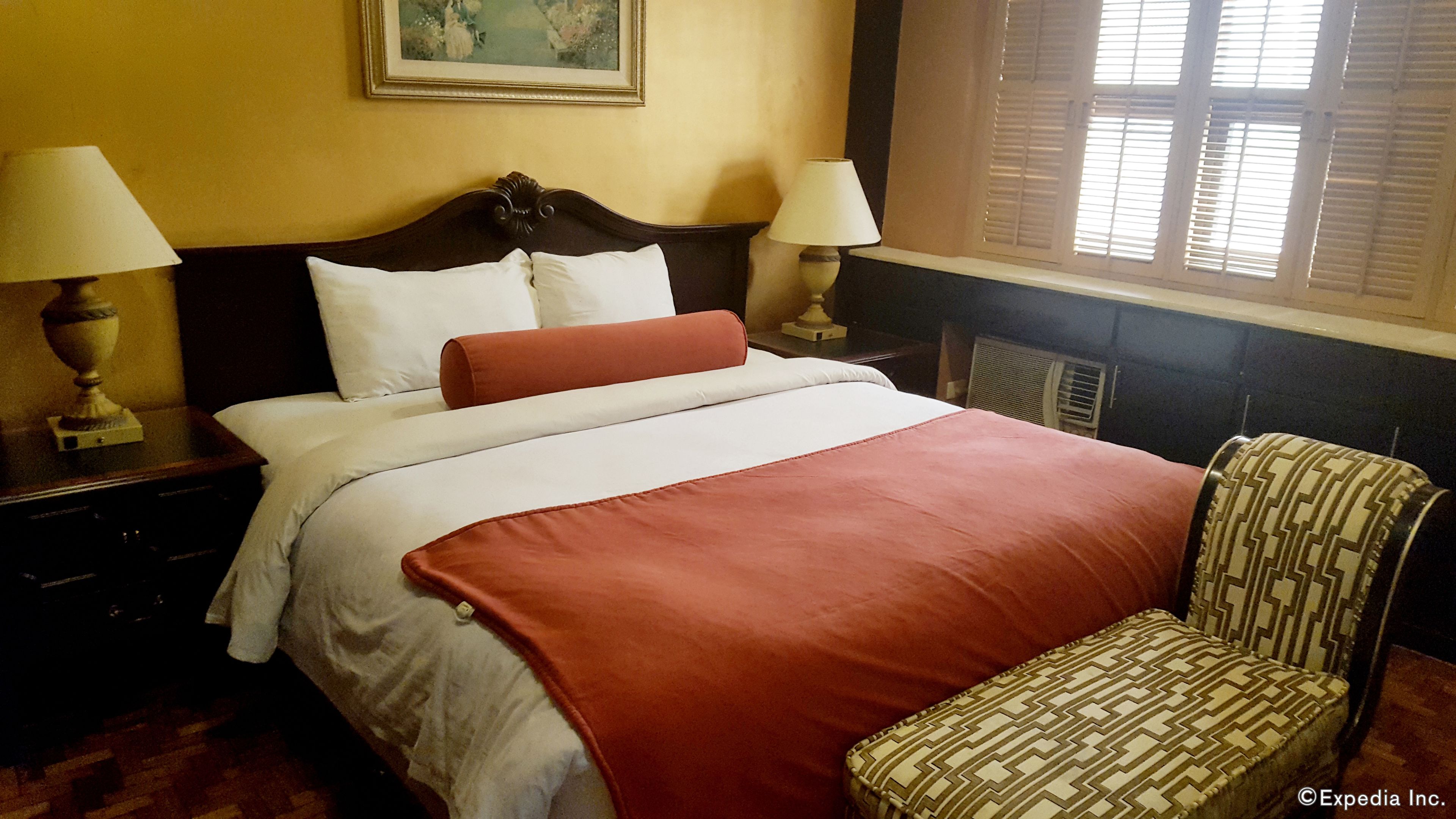 A hotel room with a large bed, two bedside lamps, a chair, and a window with shutters.