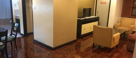 A hotel room with a wooden parquet floor, a dining area with a table and chairs, a sofa, and a TV on a cabinet.