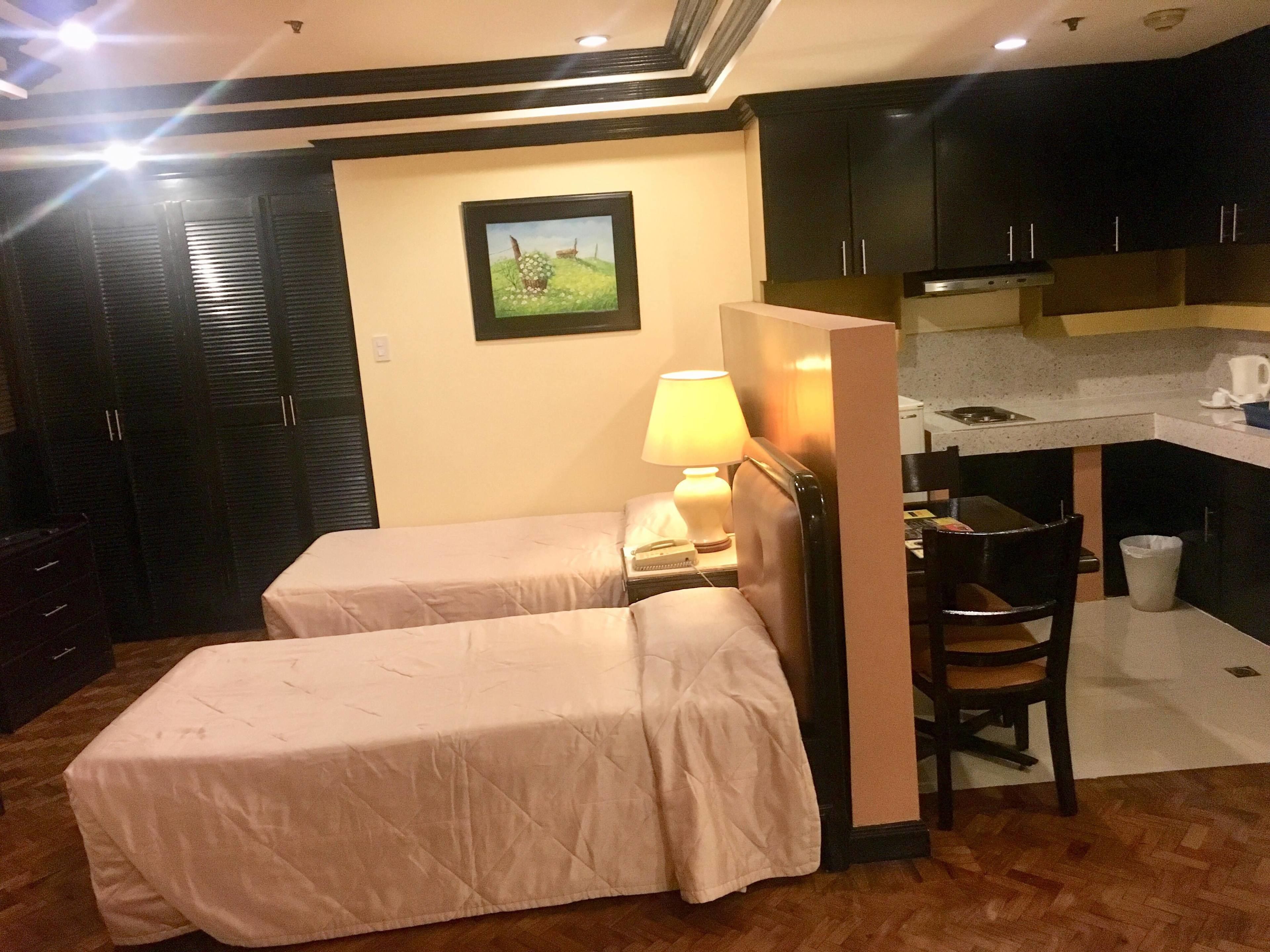 A hotel room with two beds, a desk, a chair, a lamp, and a kitchenette.