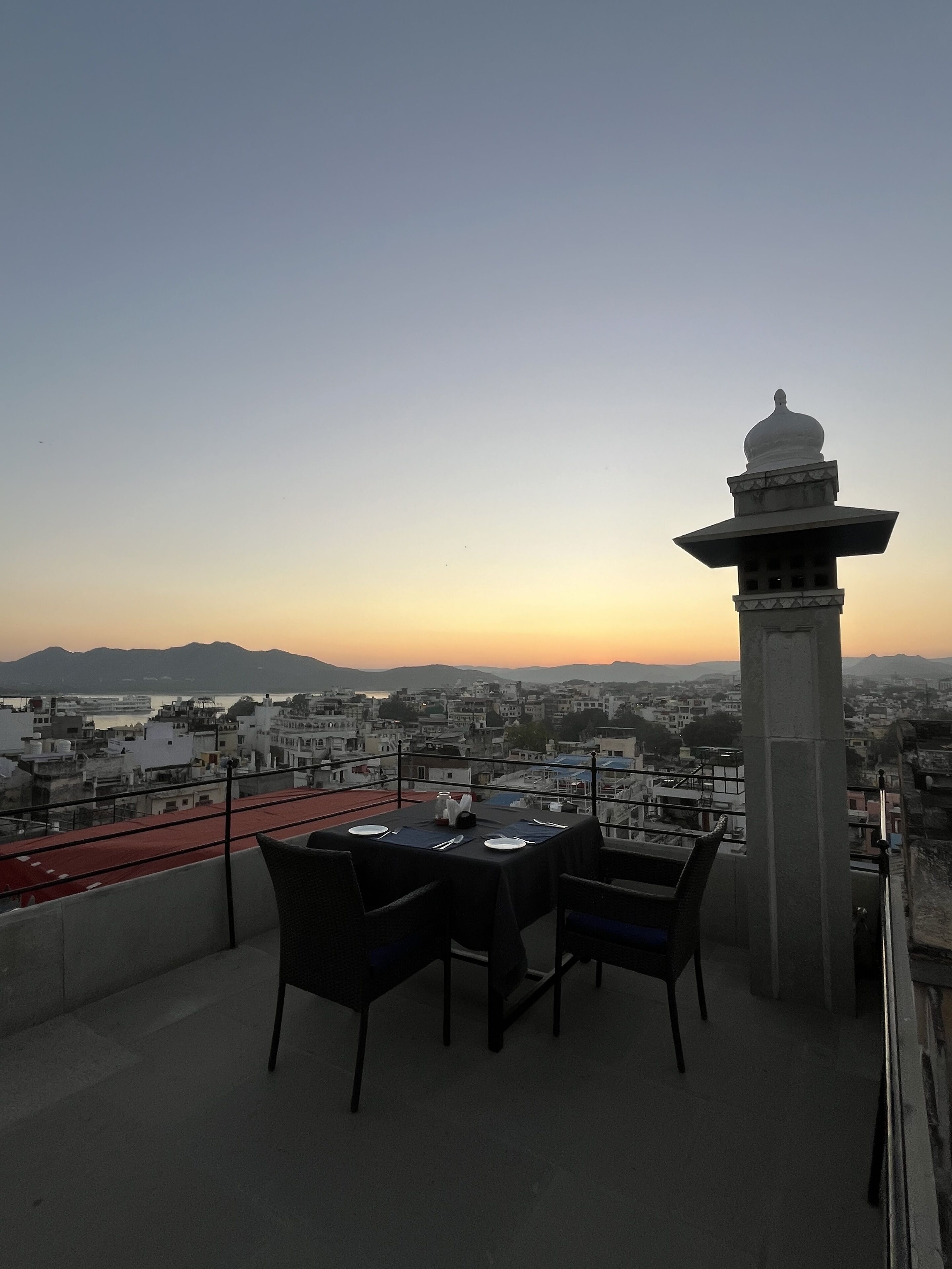 Una terraza con mesa y sillas, con vista a un paisaje urbano de edificios y una cadena montañosa distante.