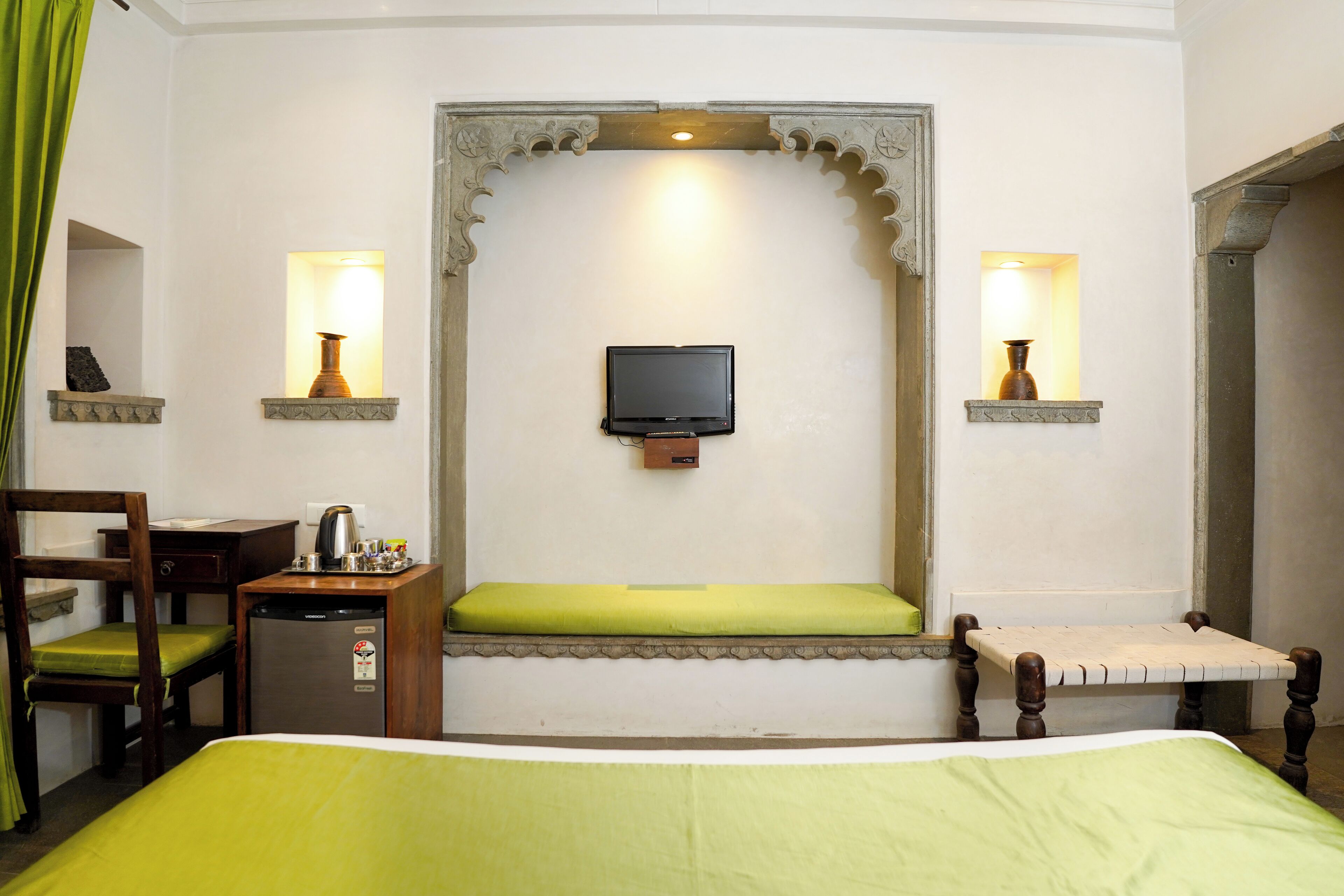 Una habitación de hotel con cama, escritorio y silla, una mesita con lámpara, televisor montado en la pared y un banco con cojín.