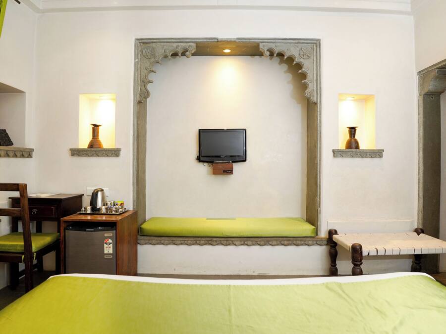 Una habitación de hotel con cama, escritorio y silla, una mesita con lámpara, televisor montado en la pared y un banco con cojín.