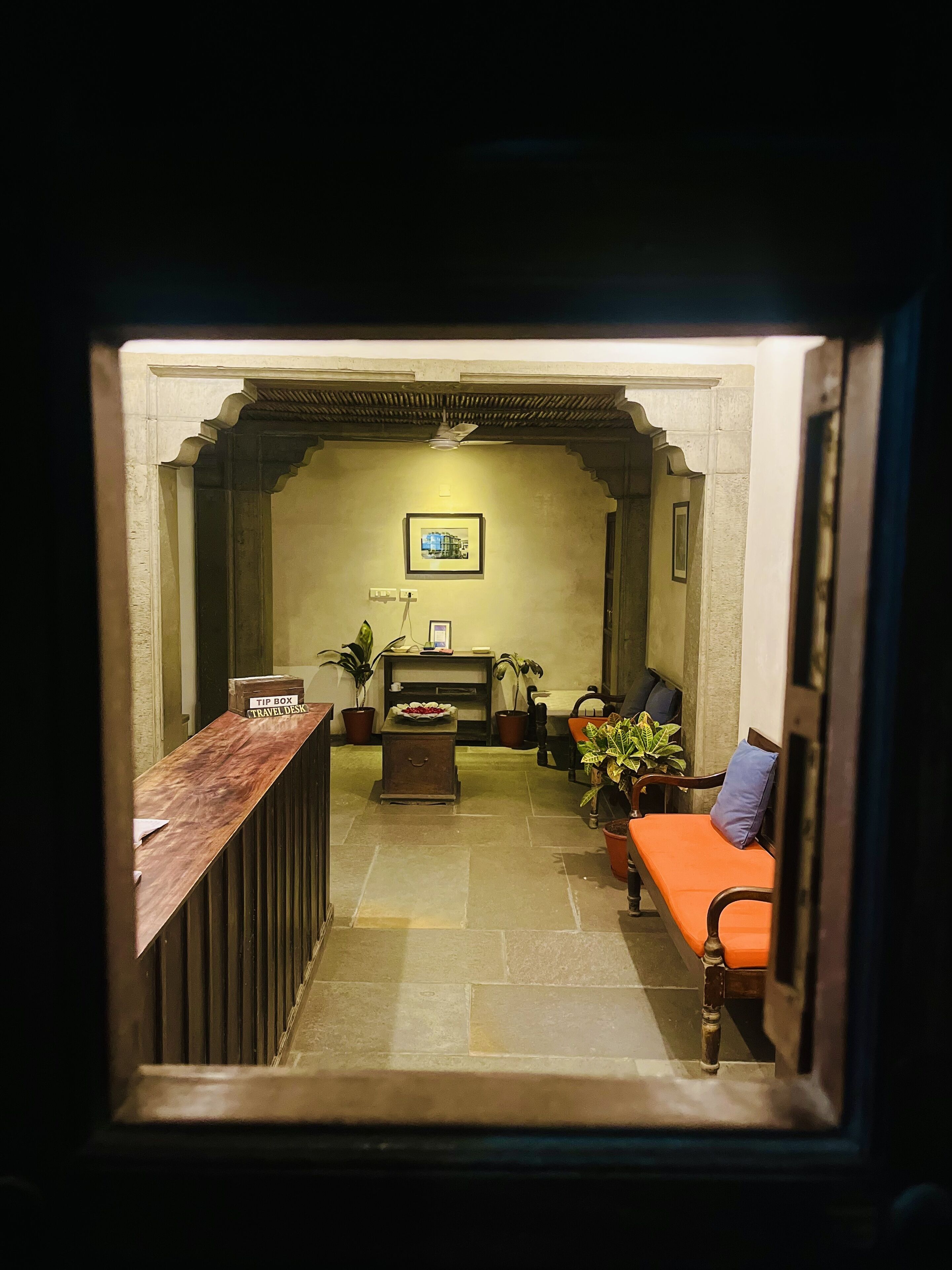 Un área de recepción con un mostrador de madera identificado con la etiqueta “TIP BOX TRAVEL DESK”, una chimenea, varias plantas en macetas y un sofá con un cojín.
