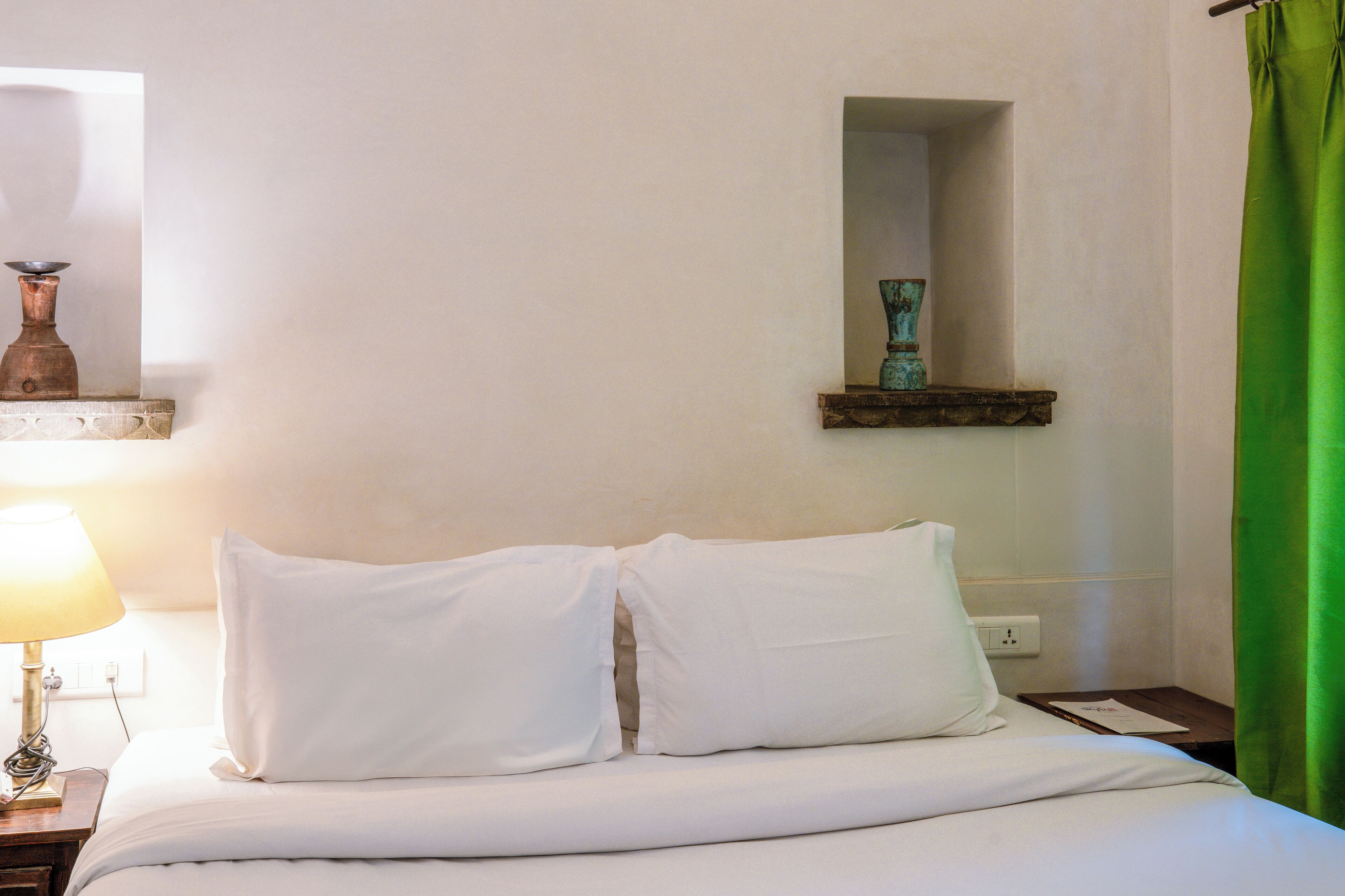 A neatly made bed with white linens, a bedside lamp, a small wooden nightstand, and a green curtain.