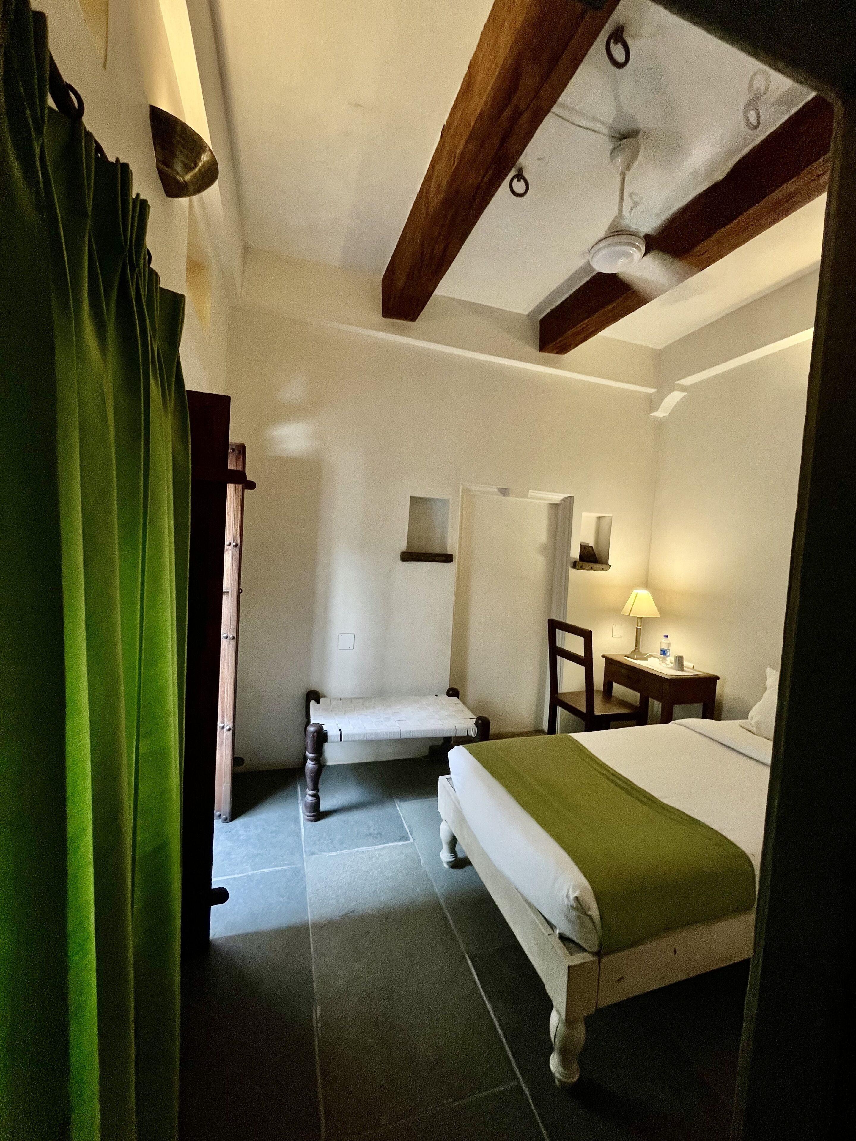 A room with a bed, a chair, a small table with a lamp, and a ceiling fan.