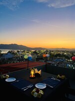 Un área de comedor en la azotea con una mesa para dos, con vistas a un paisaje urbano al atardecer.
