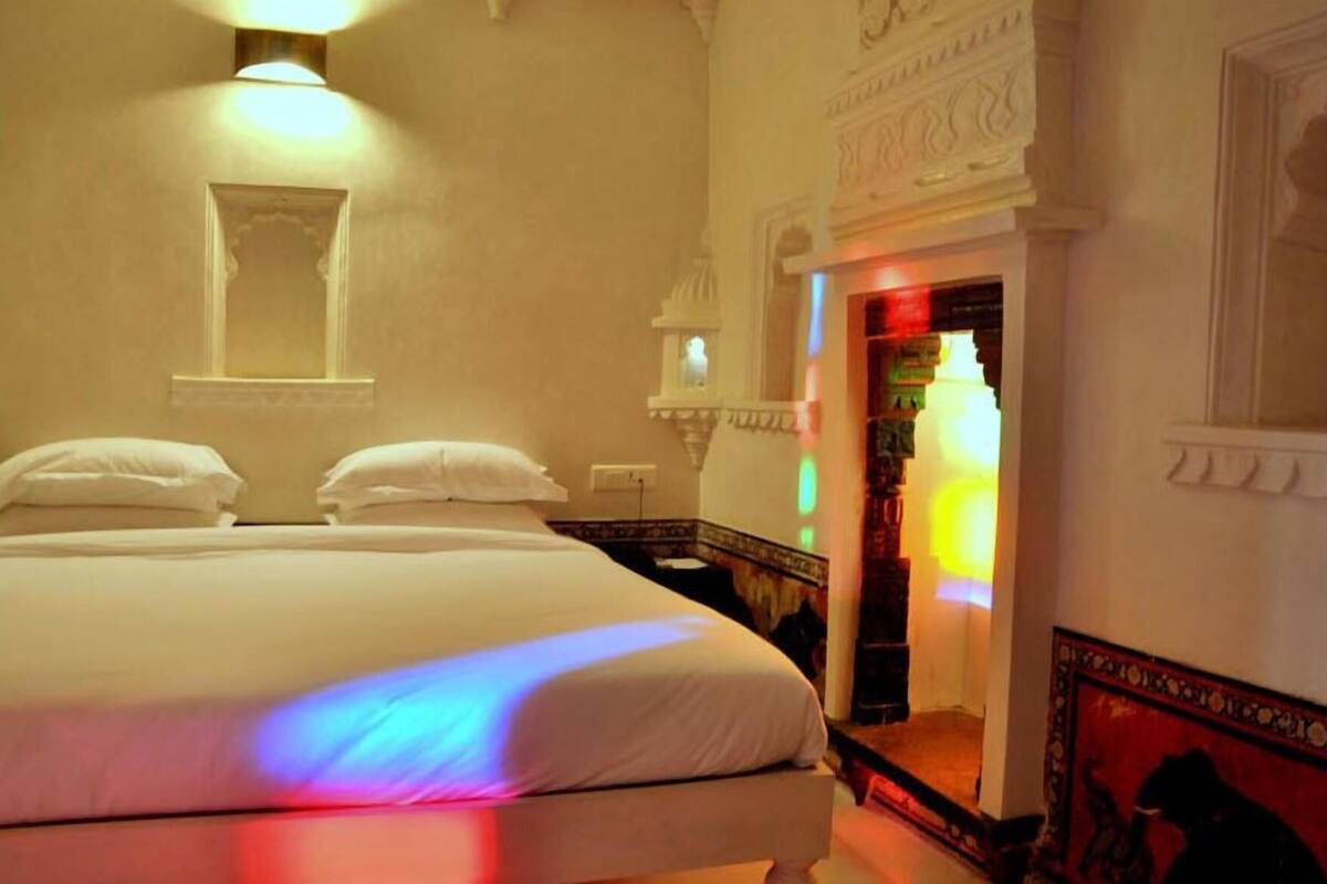 Habitació d'hotel amb un llit, una tauleta de nit, una làmpada penjada a la paret i una porta que condueix a una altra habitació.