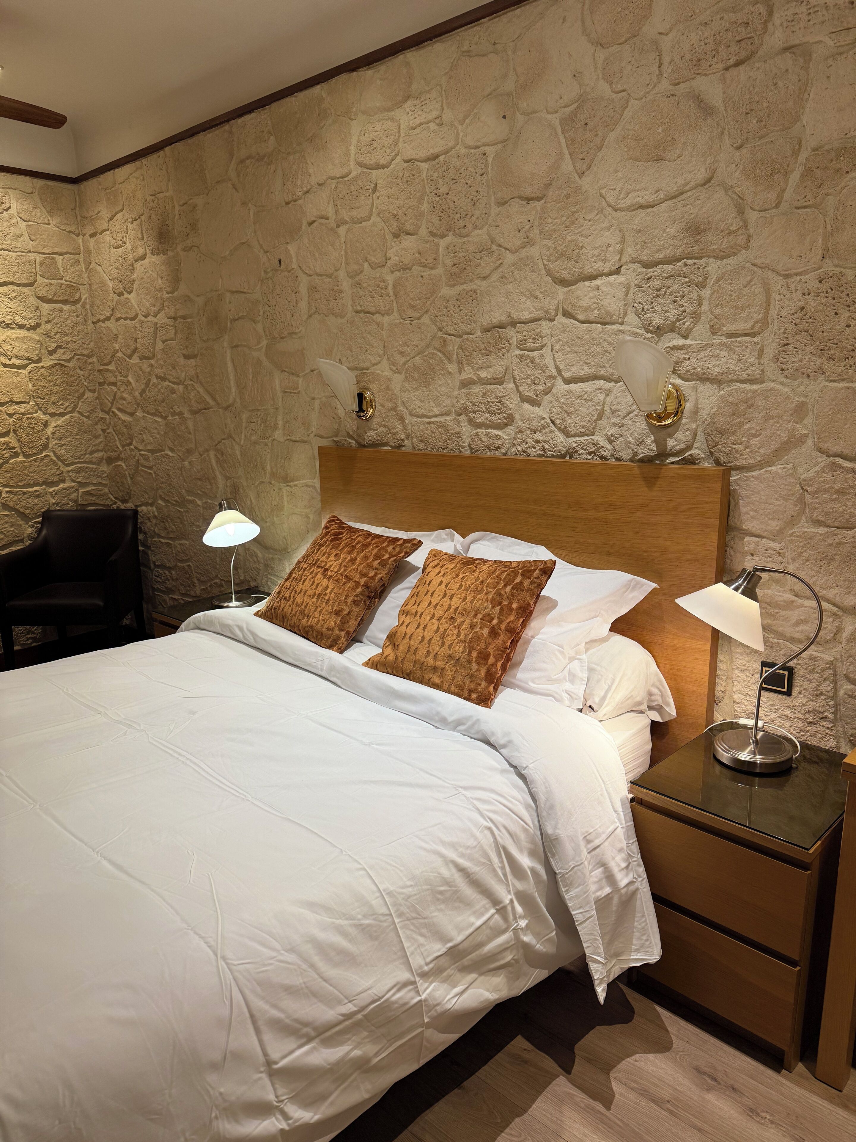 Un lit bien fait, avec du linge de lit blanc et deux coussins à motifs, une lampe de chevet et une table de chevet dotée d’un tiroir. La pièce est agrémentée d’un mur en pierre et d’une tête de lit en bois.